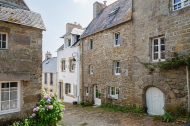 Quaint stone buildings line a sunny cobblestone alley in the historic European village of Pont-Croix, in Brittany, France. Hydrangeas bloom near a white door, ivy climbs a wall, creating a timeless, picturesque scene of rural charm.