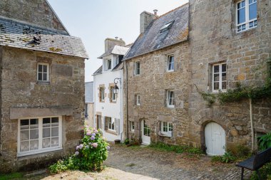 Quaint stone buildings line a sunny cobblestone alley in the historic European village of Pont-Croix, in Brittany, France. Hydrangeas bloom near a white door, ivy climbs a wall, creating a timeless, picturesque scene of rural charm.