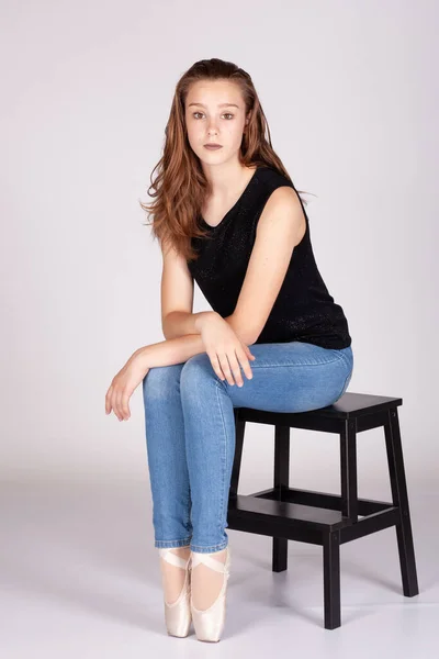 stock image En Pointe resting on stool legs together unemotional en pointe Beautiful teen girl sitting straight looking at the camera, hand to chin with an intimate straight gaze. Metaphor for trouble and secrets.