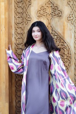 Güzel bir manken, geleneksel Özbekistan moda elbisesi içinde açık ahşap kapılı bir kız. Taşkent, Özbekistan