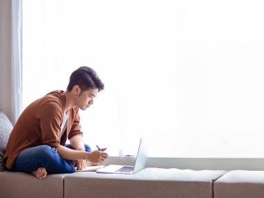 Genç Asyalı adam deftere yazı yazıyor ve dizüstü bilgisayar kullanıyor..