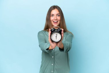 Genç ve güzel bir kadın mavi arka planda pijamalarıyla ve saatini tutarak izole edilmiş.