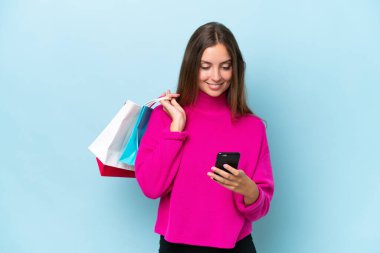 Genç ve güzel bir kadın mavi arka planda izole edilmiş. Elinde alışveriş çantaları var ve bir arkadaşına cep telefonuyla bir mesaj yazıyor.