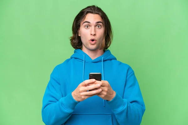 驚きの表情で携帯を使いながらカメラを見ている緑のクロマの背景に孤立した若いハンサムな男 — ストック写真