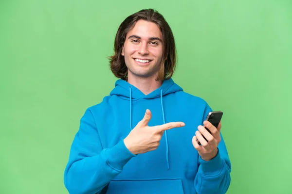年轻英俊的男子用手机在绿色的背景上隔离 并指向它 — 图库照片
