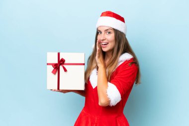 Anne Noel gibi giyinmiş, mavi arka planda izole edilmiş bir hediye taşıyan beyaz kadın bir şeyler fısıldıyor.