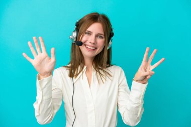 Tele pazarlamacı İngiliz kadın mavi arka planda izole edilmiş kulaklıkla çalışıyor. Parmaklarıyla sekizi sayıyor.