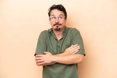 Beyazlı genç adam bej arka planda izole edilmiş. Omuzlarını kaldırırken şüpheyle hareket ediyor.