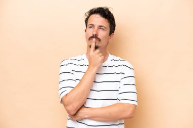 Young caucasian man isolated on beige background having doubts while looking up