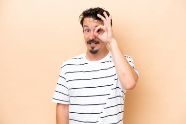 Beyazlı genç adam bej arka planda izole edilmiş. Parmaklarıyla tamam işareti gösteriyor.