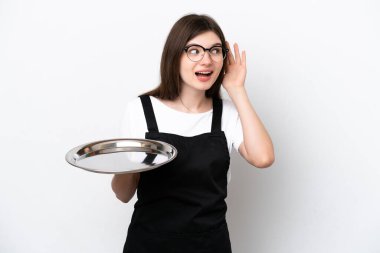 Beyaz arka planda tepsisi olan genç Rus kadın aşçı elini kulağına koyarak bir şeyler dinliyor.