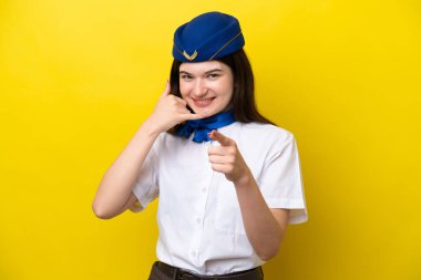 Uçak hostesi Rus kadın, sarı arka planda izole edilmiş, telefon jesti yapıyor ve ön tarafı işaret ediyor.