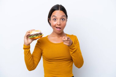 Beyaz arka planda izole edilmiş bir burger tutan genç İspanyol kadın şaşırmış ve ön tarafı işaret ediyor.
