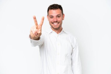 Beyaz arka planda izole edilmiş beyaz tenli yakışıklı adam gülümsüyor ve zafer işareti gösteriyor.