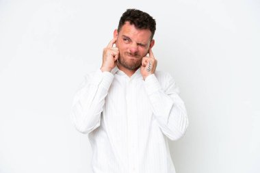 Beyaz arka planda izole edilmiş beyaz, yakışıklı genç adam hayal kırıklığına uğramış ve kulaklarını kapatmış.