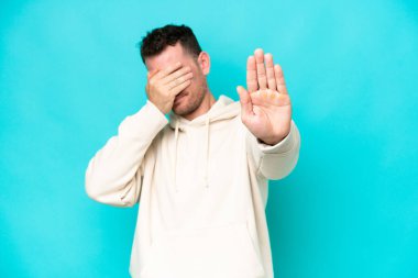 Beyaz tenli yakışıklı adam mavi arka planda izole edilmiş. Jesti durdurup yüzünü kapatıyor.