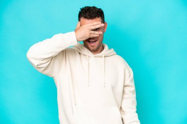 Beyaz tenli yakışıklı adam mavi arka planda izole edilmiş gözleri elleriyle kaplamış ve gülümsüyor.