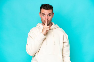 Mavi arka planda izole edilmiş beyaz, yakışıklı genç bir adam sessiz bir jest yapıyor. Parmağını ağzına sokuyor.