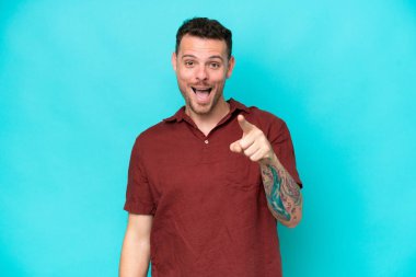 Mavi arka planda izole edilmiş beyaz, yakışıklı genç bir adam şaşırmış ve ön tarafı işaret ediyor.