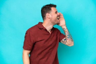 Mavi arka planda izole edilmiş, ağzı sonuna kadar açık bağıran beyaz, yakışıklı bir genç.