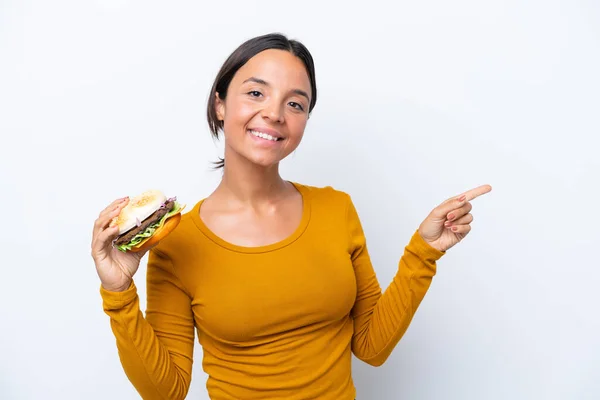 Młoda Latynoska Trzyma Hamburgera Odizolowanego Białym Tle Wskazując Palcem Bok — Zdjęcie stockowe