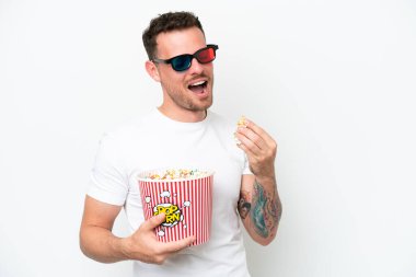 Young caucasian handsome man isolated on white background with 3d glasses and holding a big bucket of popcorns
