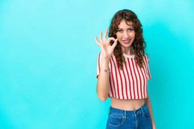 Mavi arka planda izole edilmiş kıvırcık saçlı genç kadın parmaklarıyla tamam işareti gösteriyor.
