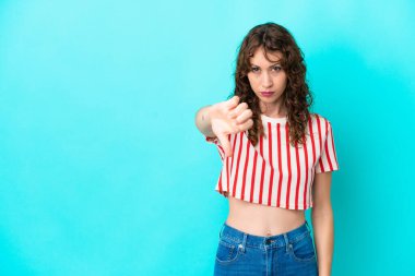 Mavi arka planda izole edilmiş kıvırcık saçlı genç bir kadın olumsuz bir ifadeyle baş parmağını aşağı gösteriyor.