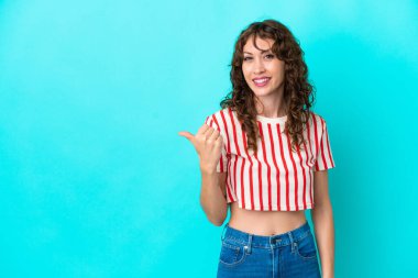 Mavi arka planda izole edilmiş kıvırcık saçlı genç bir kadın bir ürünü sunmak için kenarı işaret ediyor.