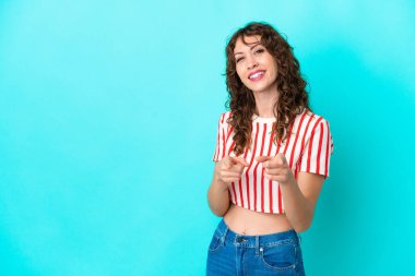 Mavi arka planda izole edilmiş kıvırcık saçlı genç kadın şaşırmış ve ön tarafı işaret ediyor.