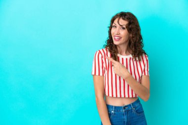 Mavi arka planda yalıtılmış kıvırcık saçlı genç kadın şaşırmış ve işaret ediyor.