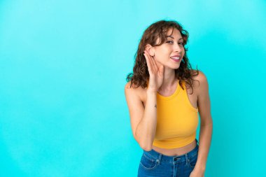 Mavi arka planda izole edilmiş kıvırcık saçlı genç bir kadın elini kulağına koyarak bir şeyler dinliyor.