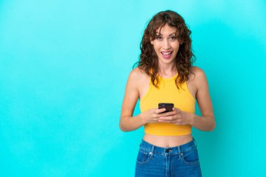 Mavi arka planda izole edilmiş kıvırcık saçlı genç kadın şaşırdı ve bir mesaj gönderiyor.