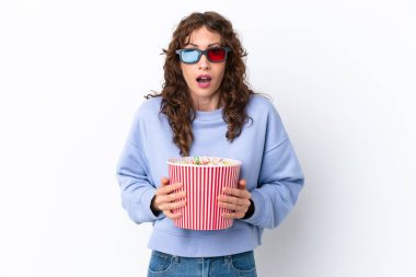 Young woman with curly hair isolated on white background surprised with 3d glasses and holding a big bucket of popcorns