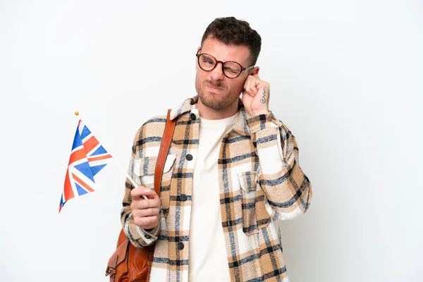 Young hispanic man holding an United Kingdom flag isolated on white background frustrated and covering ears