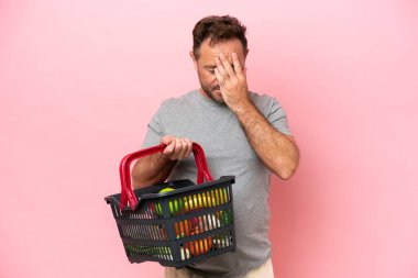 Elinde alışveriş sepeti tutan orta yaşlı beyaz bir adam. Pembe arka planda, yorgun ve hasta bir ifadeyle izole edilmiş.