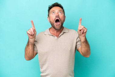 Mavi arka planda izole edilmiş orta yaşlı beyaz adam şaşırmış ve işaret ediyor.