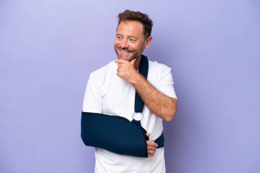 Kırık kollu, mor arka planda izole edilmiş askısı olan orta yaşlı beyaz bir adam yana bakıyor ve gülümsüyor.