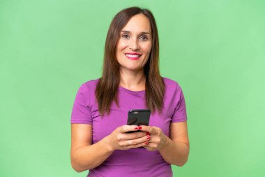 Orta yaşlı beyaz kadın izole edilmiş arka planda kameraya bakıyor ve cep telefonunu kullanırken gülümsüyor.