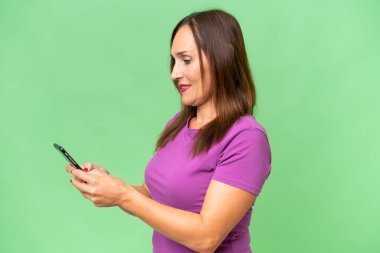 Orta yaşlı beyaz kadın izole edilmiş arka planda cep telefonuyla mesaj ya da e-posta gönderiyor.