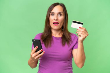 Orta yaşlı beyaz bir kadın cep telefonuyla alışveriş yapıyor ve elinde şaşırmış bir ifadeyle bir kredi kartı tutuyor.