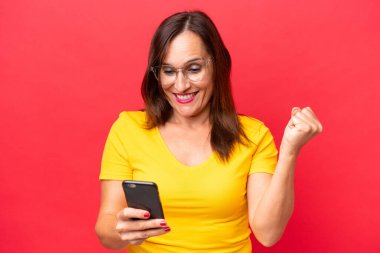 Orta yaşlı beyaz kadın, kırmızı arka planda izole edilmiş, şaşırmış ve bir mesaj gönderiyor.