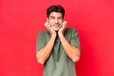 Kızıl arka planda izole edilmiş genç beyaz yakışıklı adam hayal kırıklığına uğramış ve kulaklarını kapatmış.