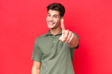 Genç, beyaz, yakışıklı bir adam kırmızı arka planda izole edilmiş ve parmağını kaldırıyor.