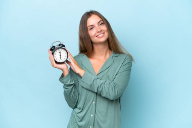 Genç ve güzel bir kadın mavi arka planda pijamalarıyla ve mutlu bir ifadeyle saati tutuyor.