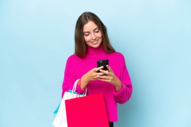 Genç ve güzel bir kadın mavi arka planda izole edilmiş. Elinde alışveriş çantaları var ve bir arkadaşına cep telefonuyla bir mesaj yazıyor.