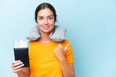 Şişirilebilir seyahat yastığı olan genç ve güzel bir kadın mavi arka planda izole edilmiş bir şekilde bir ürün sunmak için kenarı işaret ediyor.