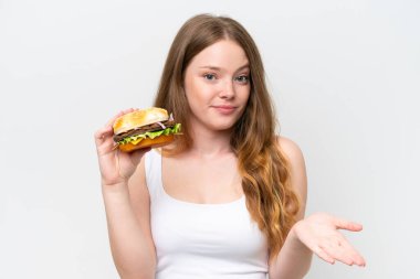Genç, güzel bir kadın, beyaz arka planda izole edilmiş bir burger tutuyor. Omuzlarını kaldırırken şüphe uyandırıyor.