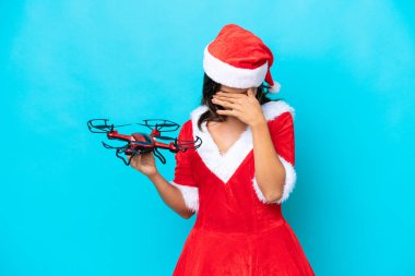 Anne Noel gibi giyinmiş genç bir kadın elinde mavi arka planda, yorgun ve hasta bir ifadeyle izole edilmiş bir dron tutuyor.