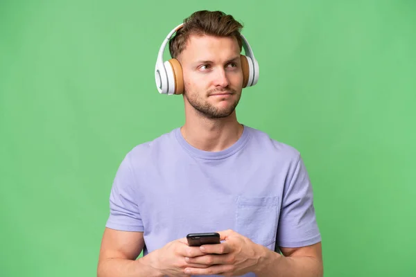 年轻的金发碧眼的白人男子在孤独的背景下一边用手机一边听音乐一边思考 — 图库照片